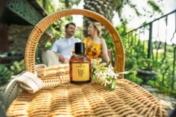 Güneşli hasır sepet üzerinde Harmony parfüm şişesi, arka planda yasemin ve bulanık çift, Avrupa'da yerinde cilalı ürün fotoğrafçılığı