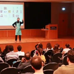 Fotograf, der auf der Bühne ein praktisches Fotoseminar leitet, mit blaugrüner Schürze und Diaprojektor im Rücken, unterrichtet in Berlin.