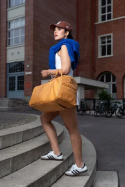 Berlin'de yerinde moda çekimi: taba rengi deri spor çantası, mavi kazağı ve şapkasıyla basamakları tırmanan kadın, sinematik sokak portresi.