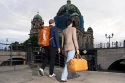 Bulutlu bir gökyüzünün altında, taş bir köprüden geçerek kubbeli tarihi katedrale doğru yürüyen, arkadan görülen iki kişi büyük kahverengi deri seyahat çantaları taşıyor — Fotoğraf: Burak Bulut Yıldırım; anahtar kelimeler: seyahat, kentsel, mimari, yaşam tarzı, deri