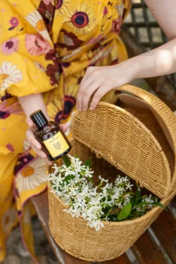 Eller, sarı etiketli bir kokulu yağ şişesini beyaz çiçeklerden oluşan dokuma bir sepetin içine yerleştiriyor - Avrupa için yerinde ürün fotoğrafçılığı.