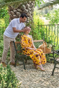 Bir adam bahçede oturan bir kadının gözlerini kapatıyor; kadın sarı çiçekli bir elbise giyiyor - Avrupa için yerinde sinematik moda çekimi