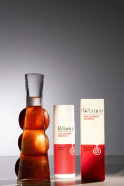 Rélance kepek önleyici şampuan şişesi ve kutusunun yanında turunçgillerle dolu, cilalı kehribar renkli bir bardağın Berlin'de stüdyo çekimi ürün fotoğrafı.