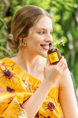 Parlak ayçiçeği desenli elbise giymiş kadın, bulanık yeşil bahçe fonunda küçük beyaz çiçeklerle süslenmiş Harmony yazılı kehribar rengi parfüm şişesini tutarken gülümsüyor; Fotoğraf: Burak Bulut Yıldırım — canlı, doğal, yaşam tarzı, renkli, çiçekli