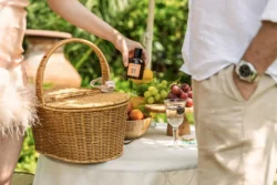 Beyaz örtülü bir açık hava masasında, meyve kaseleri ve kırpılmış bir çiftin bulunduğu hasır piknik sepeti, bir eliyle üzümlerin arasına küçük bir kehribar şişe yerleştirmeye çalışan bir çift; Fotoğraf: Burak Bulut Yıldırım; Anahtar kelimeler: piknik, yaşam tarzı, yaz, rustik, doğal