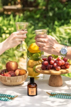 Bir parfüm şişesi, meyve ve üzümler üzerinde kızaran iki elin güneşli yaşam tarzı ürün çekimi - Avrupa'da gösterişli ürün fotoğrafçılığı.