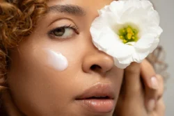 Nemli cildi, parlatılmış dudakları ve bir gözünün üzerinde tuttuğu beyaz çiçeği ile yanağında bir tutam krem bulunan bir kadının yakın çekim portresi; Fotoğraf: Burak Bulut Yıldırım; anahtar kelimeler: yakın çekim, doğal, cilt bakımı, çiçek, nemli