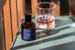 Yeşil kumaş üzerinde bir viski bardağının yanında Alpha etiketli kehribar cam parfüm şişesi, sıcak güneş ışığı; Berlin'de cilalı ürün fotoğrafçılığı.