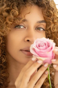 Yumuşak doğal ışıkta pembe bir gül tutan kıvırcık saçlı bir kadının yakın çekim güzellik portresi, Avrupa çapında cilalı güzellik fotoğrafçılığı.