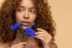 Yumuşak stüdyo ışığında kıvırcık saçlı model bej bir arka plan üzerinde canlı mavi çiçekler tutuyor - Avrupa'daki projeler için gösterişli güzellik fotoğrafçılığı.