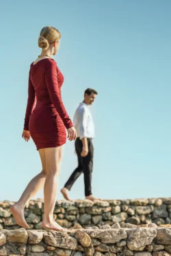 Kırmızı bir elbise giymiş çıplak ayaklı model, arkasında bulanık bir adamla güneşin aydınlattığı bir taş duvarda yürüyor - sinematik yerinde moda çekimi, Avrupa.