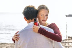 Güneşli bir plajda bir erkeği kucaklayan ve elinde parfüm şişesi tutan bir kadının cilalı güzellik ürünü fotoğrafı, Avrupa'daki projeler için fotoğrafçı.