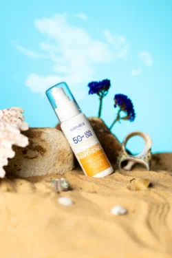 Deniz kabukları ve mavi fon ile kumda açılı bir güneş kremi pompası şişesinin cilalı ürün fotoğrafçılığı, Avrupa'daki projeler için fotoğrafçı.