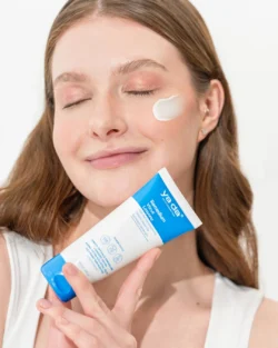 Yanağında krem, elinde mavi-beyaz bir tüp tutan gülümseyen kadın; Berlinli bir fotoğrafçıdan Avrupa için cilalı güzellik ürünleri fotoğrafçılığı.