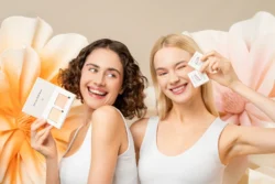 Gülümseyen iki model, yumuşak çiçekli fonda cilt bakımı numuneleri tutuyor, Avrupa'daki projeler için cilalı güzellik ve ürün fotoğrafçılığı.