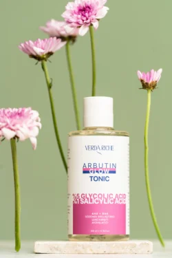 Yumuşak stüdyo aydınlatması ve pembe çiçeklerle taş levha üzerinde Arbutin Glow tonik şişesinin cilalı ürün fotoğrafçılığı, Avrupa'daki projeler için fotoğrafçı.