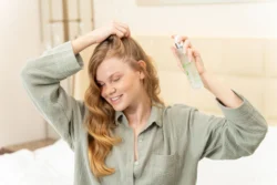 Yumuşak ışıkta gülümseyen kızıl saçlı, yatağın üzerinde buzlu bir saç buğusu şişesini püskürtür - Berlin'de cilalı güzellik ürünü çekimi.