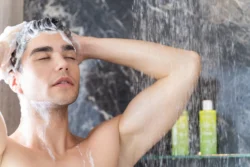 Şampuan köpüğüyle gözleri kapalı bir şekilde akan duşun altında saçını yıkayan adam, cam rafta yeşil şampuan şişeleri ve arka planda koyu renkli mermer fayanslar; Fotoğraf: Burak Bulut Yıldırım — rahatlatıcı, temiz, samimi, ferah, yaşam tarzı.