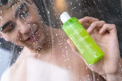 Sabunlu duş camının arkasında parlak yeşil biberiye şampuanı şişesini tutan bir modelin cilalı ürün fotoğrafı, Avrupa'da fotoğrafçı.