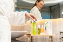 Beyaz bornozlu kadın, banyo lavabosunun yanındaki ahşap tepside duran yeşil renkli cilt bakım ürününü almaya uzanırken, aynada gülümseyen yansıması görülüyor — Fotoğraf: Burak Bulut Yıldırım; anahtar kelimeler: cilt bakımı, güzellik, yaşam tarzı, doğal, spa