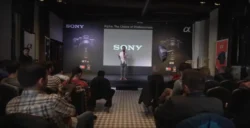 Karanlık bir sahnedeki sunucu, oturan konuklara Sony Alpha ürün konuşması yapıyor, karamsar aydınlatma, Berlin'de uygulamalı fotoğrafçılık semineri.